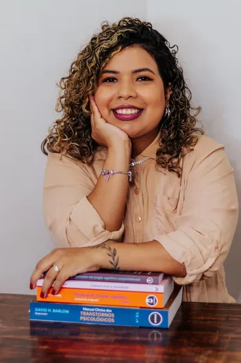 Stéfanie Miranda Barroso, psicóloga clínica, sorrindo com o rosto apoiado na mão, ao lado de uma pilha de livros de psicologia