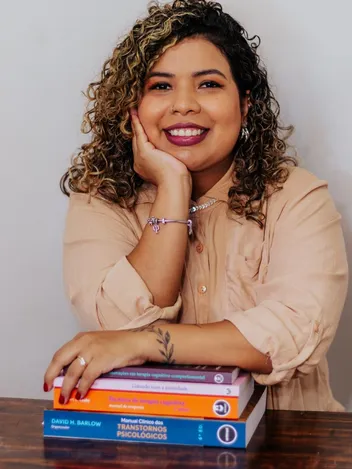 Stéfanie Miranda Barroso, psicóloga clínica, sorrindo com o rosto apoiado na mão, ao lado de uma pilha de livros de psicologia